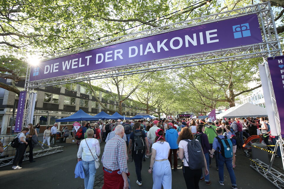 Menschen gehen unter einer Traverse hindurch. Auf der Traverse steht in weißen Buchstaben auf lila Hintergrund "Die Welt der Diakonie"