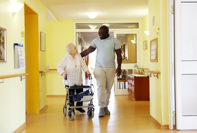 Ein Pfleger geht mit einer älteren Dame einen Gang im Altenheim entlang. Sie schauen sich freundlich an