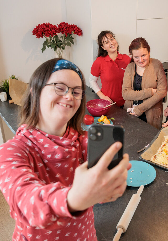 Eine junge Frau steht in der Küche ihrer Wohngruppe. Sie macht ein Selfie mit anderen jungen Menschen
