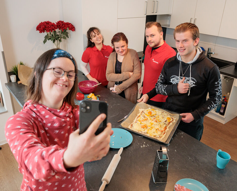 Eine junge Frau steht in der Küche ihrer Wohngruppe. Sie macht ein Selfie mit anderen jungen Menschen