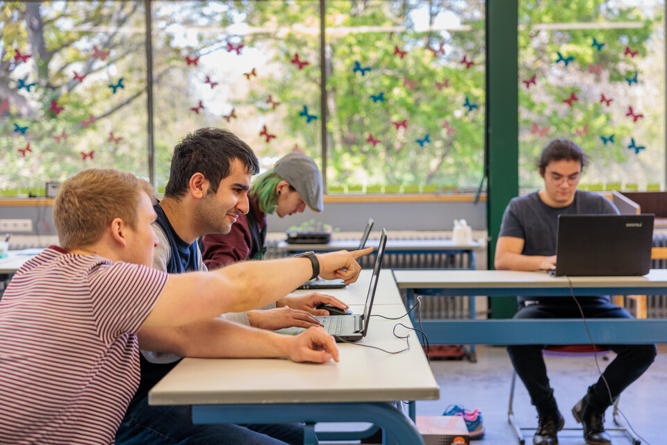 Junge Menschen sitzen in einem Schulraum, einer zeigt einem anderen etwas auf dem Laptop