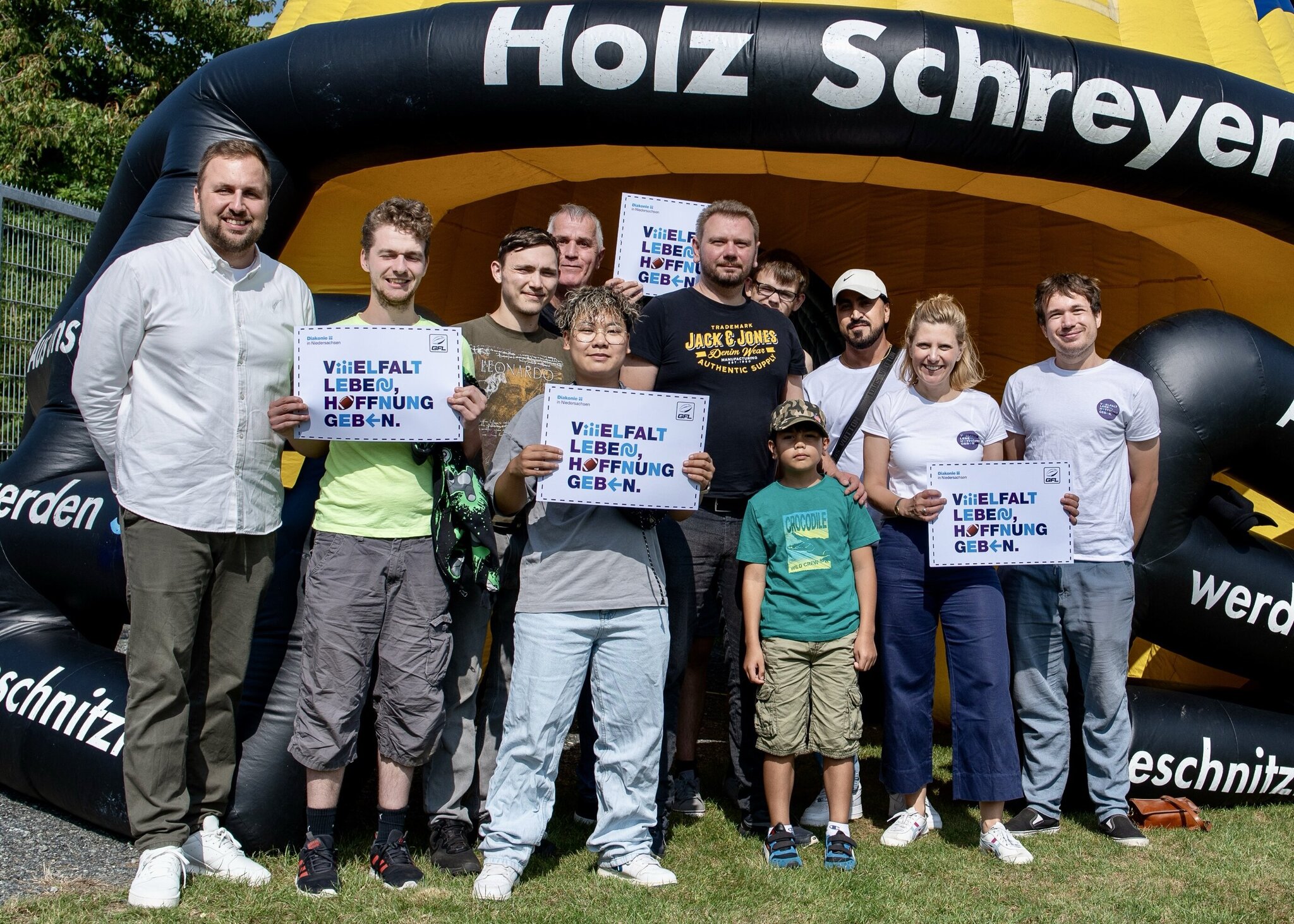 Mehrere Menschen stehen unter einem Pavillon nebeneinander. Sie schauen fröhlich in die Kamera, einige von ihnen halten Schilder mit der Aufschrift "Vielfalt leben, Hoffnung geben" in die Kamera