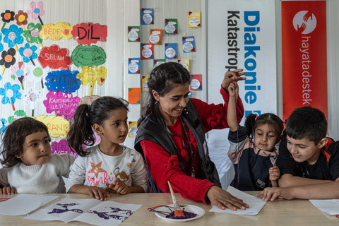 Eine Frau spielt mit mehreren Kindern. Hinter ihr ist das Banner der Diakonie Katastrophenhilfe zu sehen